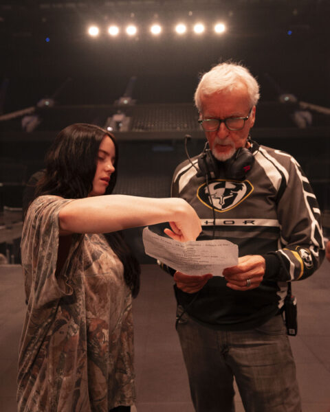 Directors Billie Eilish and James Cameron stand together looking at a piece of paper.